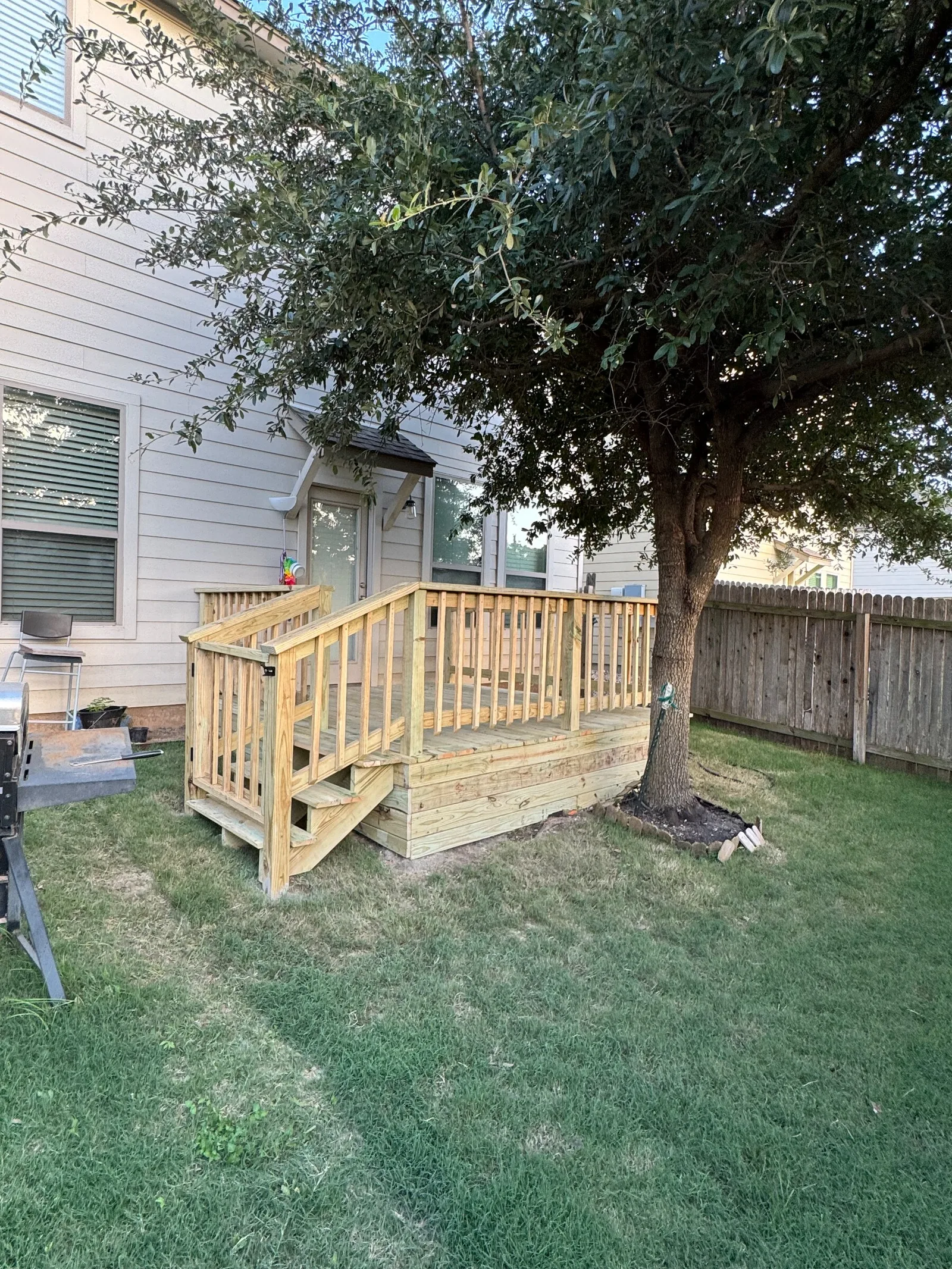 A small wood porch with railings and steps beside a house beneath a shade tree.